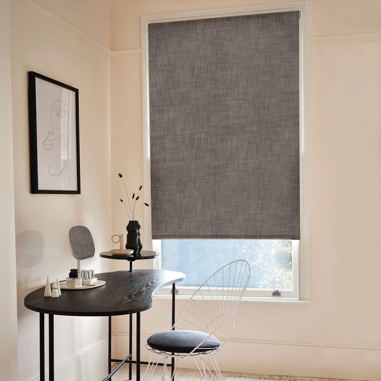 Gray textured roller blind, fully lowered and filtering light; home office with black curved desk, wire chair, minimalist line-art on wall, vase and mirror, soft daylight from lower window.