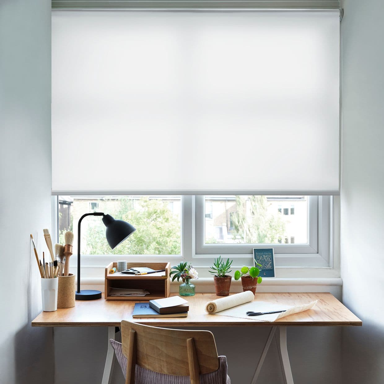 White roller blind lowered almost fully across the window, opaque fabric softly diffusing light; filtering daylight over a tidy wooden desk with lamp, brushes, small potted plants and suburban view.