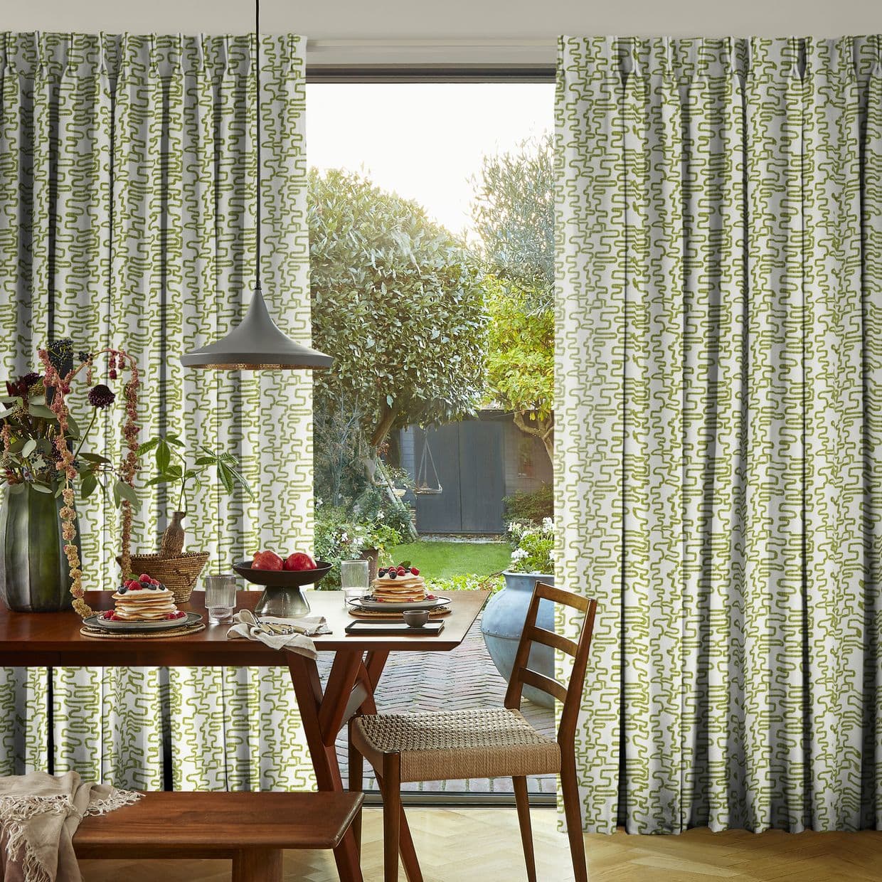 Full-length patterned curtains (green abstract squiggles) on a track, parted centrally; framing a sliding glass door and admitting soft daylight into a dining area set for breakfast with a garden view.