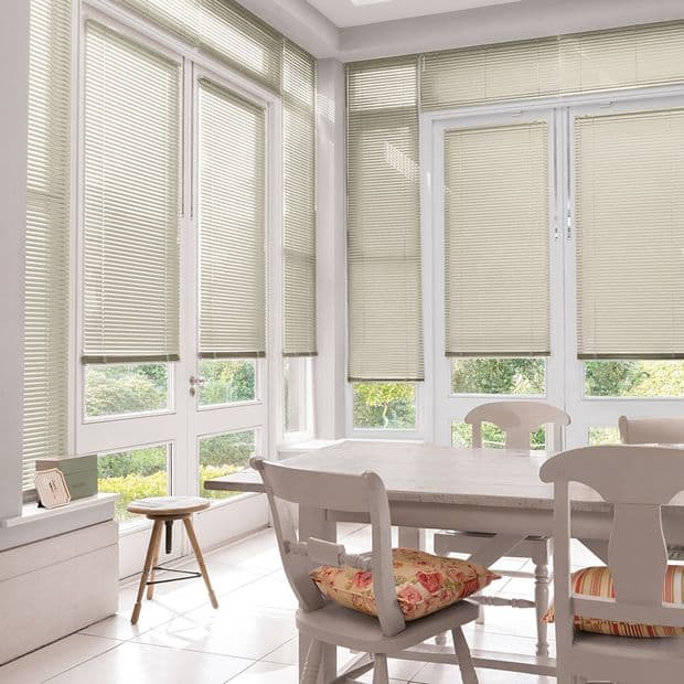 Horizontal Venetian blinds on tall bay windows and glass doors, fully lowered and gently filtering daylight; bright dining/sunroom with white wooden table and chairs, floral cushions, and green garden view.