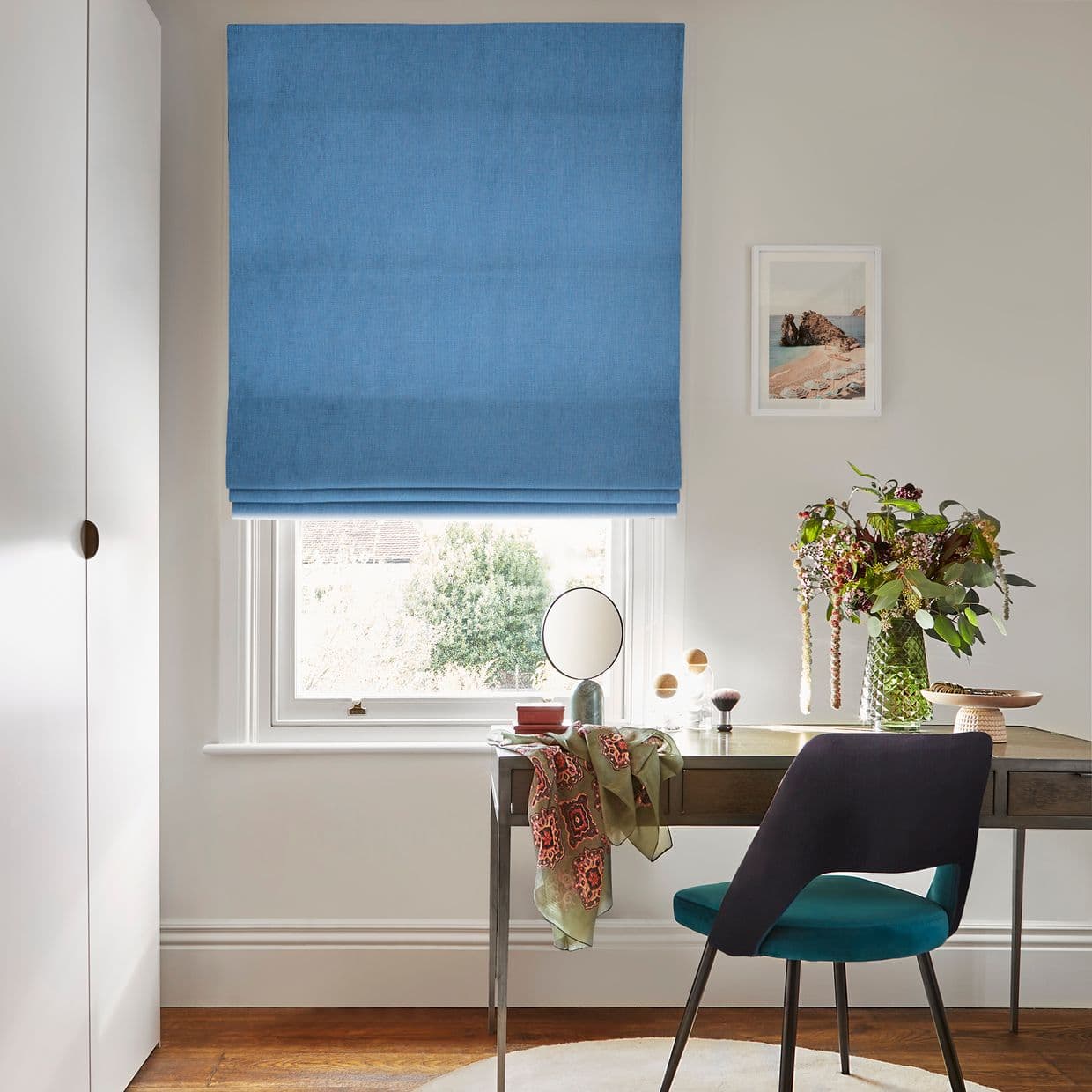 Blue Roman blind, lowered with horizontal folds, dimming incoming light; mounted over a white sash window above a wooden desk holding a chair, small mirror and a vase of flowers.