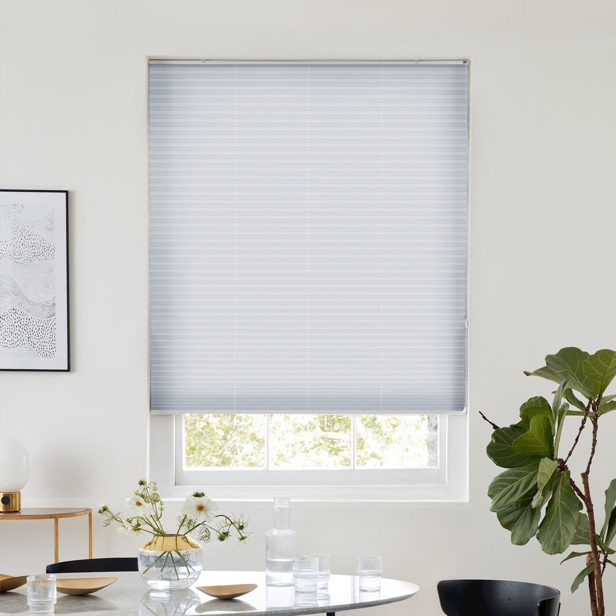 Light-gray cellular blind, lowered with a narrow bottom gap, filtering soft daylight; over a sash window in a bright dining nook with a round table, glass vase, dishes, and a fiddle-leaf fig.