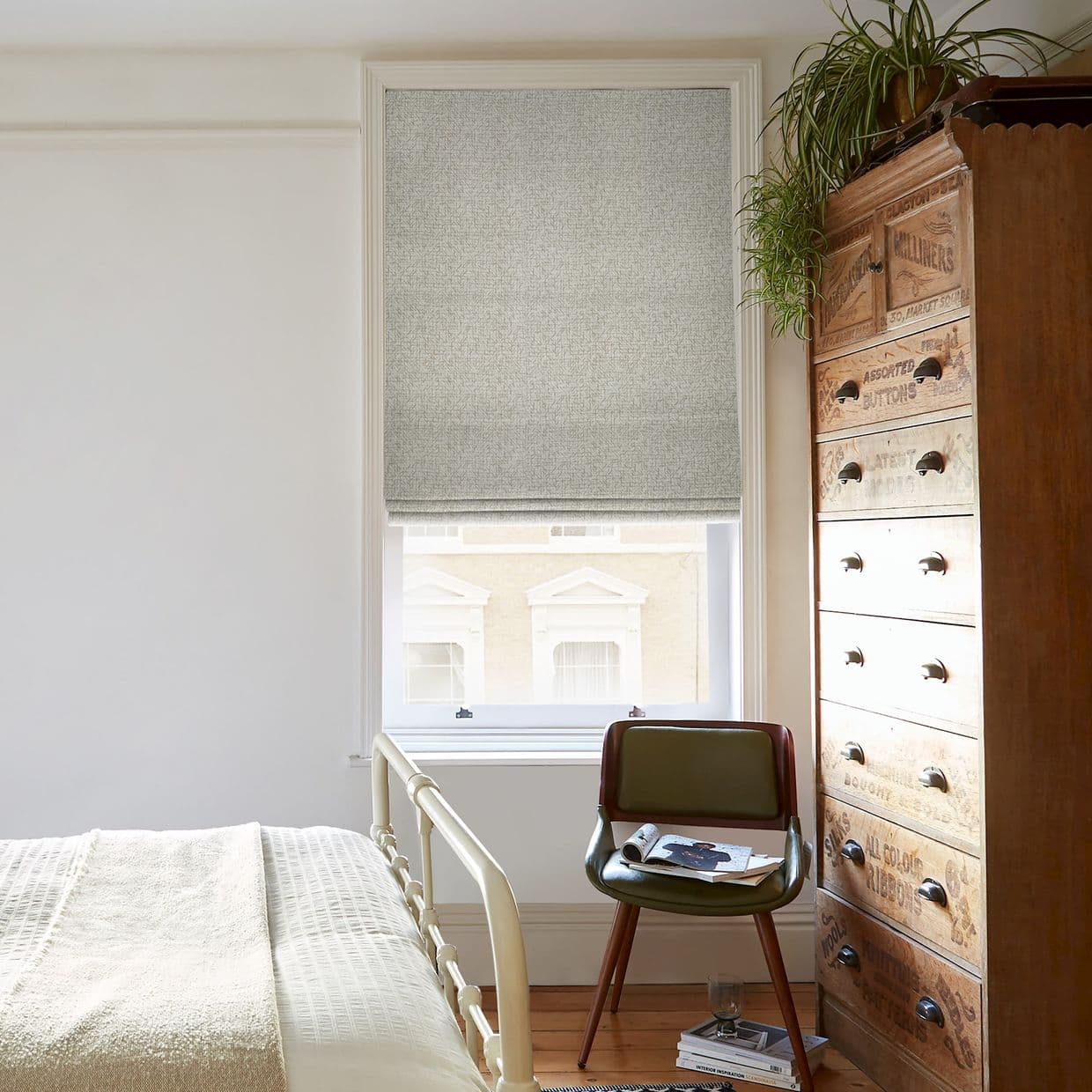 Roman fabric Roman blind, lowered, textured grey, filtering soft daylight into a cozy bedroom with a pale bed, green chair, tall vintage wooden chest, and a hanging spider plant.

ASSORTED BUTTONS; ALL COLOUR BUTTONS