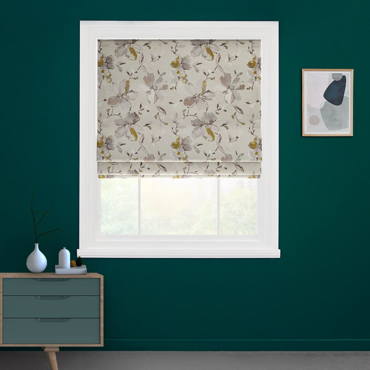 Floral Roman blind, fully lowered with folds and beige, mustard and gray blossoms, filtering daylight through a narrow lower gap; mounted in a white-framed window in a teal-walled room.