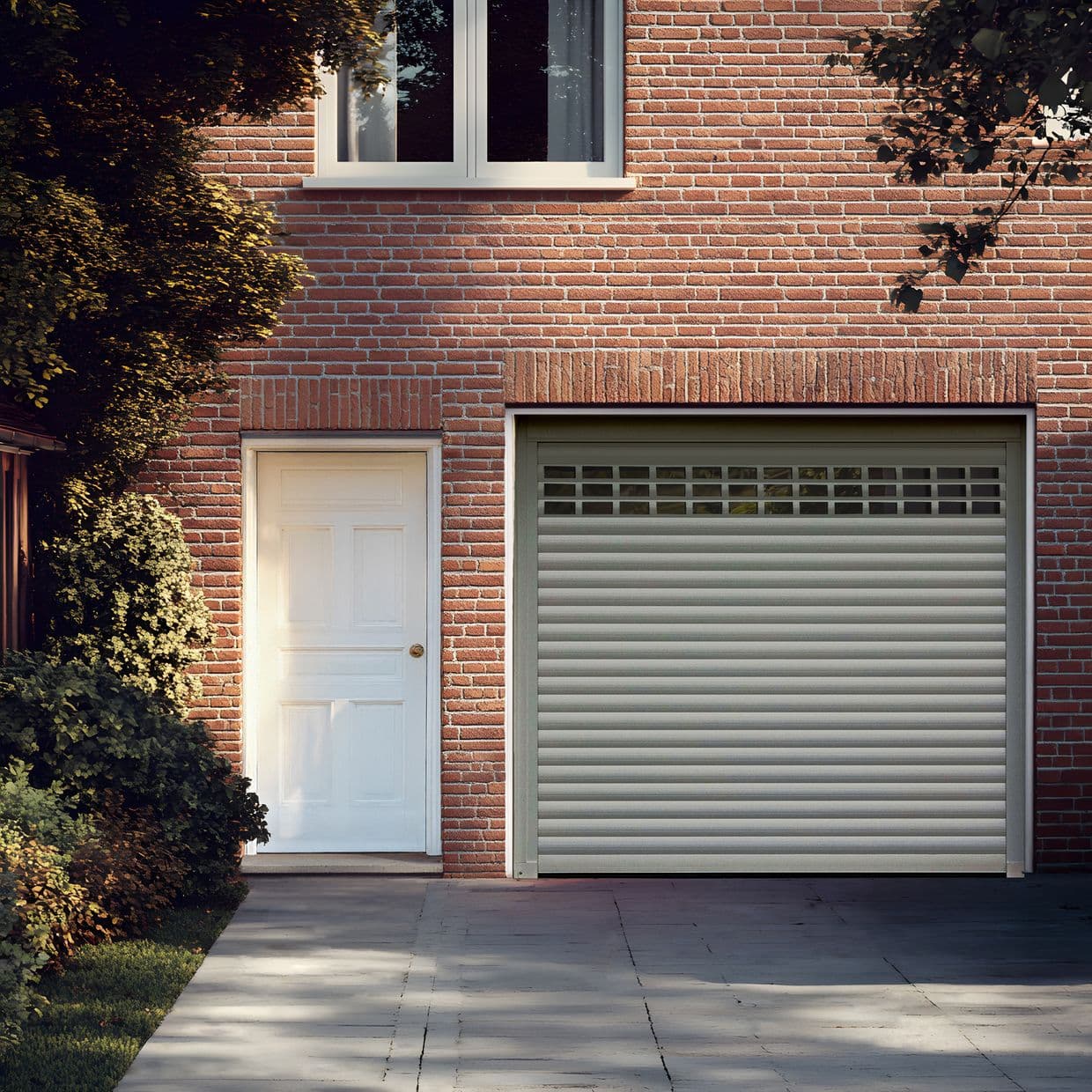 Sheer white curtains (upper window) â€” closed, softly diffusing daylight â€” against a red brick house faÃ§ade with a white side door, closed roll-up garage door, driveway and leafy shrubs.