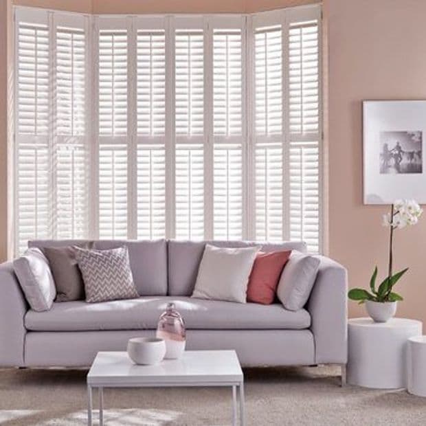 White full-height plantation shutters across a multi-panel bay window, closed with slats filtering bright daylight; behind a pale gray sofa with cushions, coffee table, orchid, and soft pink walls.