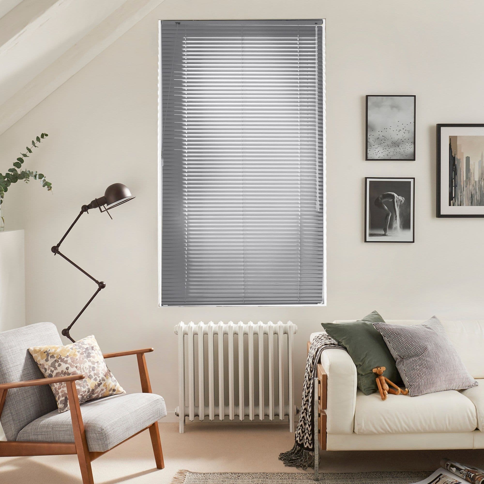 Horizontal aluminum Venetian blinds covering a tall central window, closed and gently filtering daylight; set above a radiator in a neutral-toned living room with armchair, lamp, sofa, and framed art.