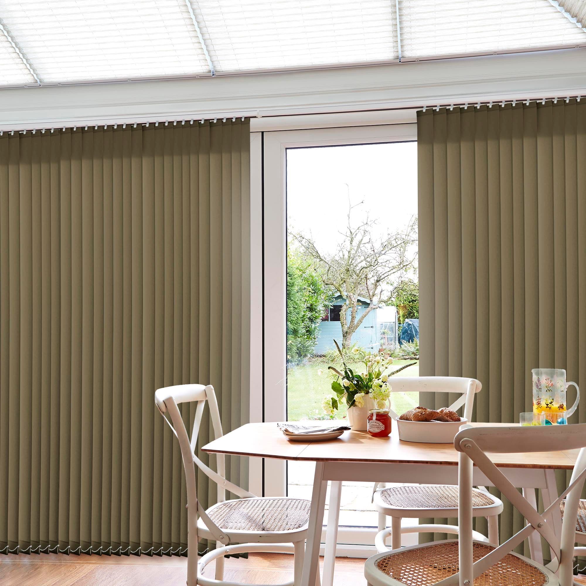 Beige vertical blinds, full-length slats mostly closed but parted centrally over a glass patio door, filtering daylight; small wooden dining table and chairs inside, soft light, garden and shed visible.