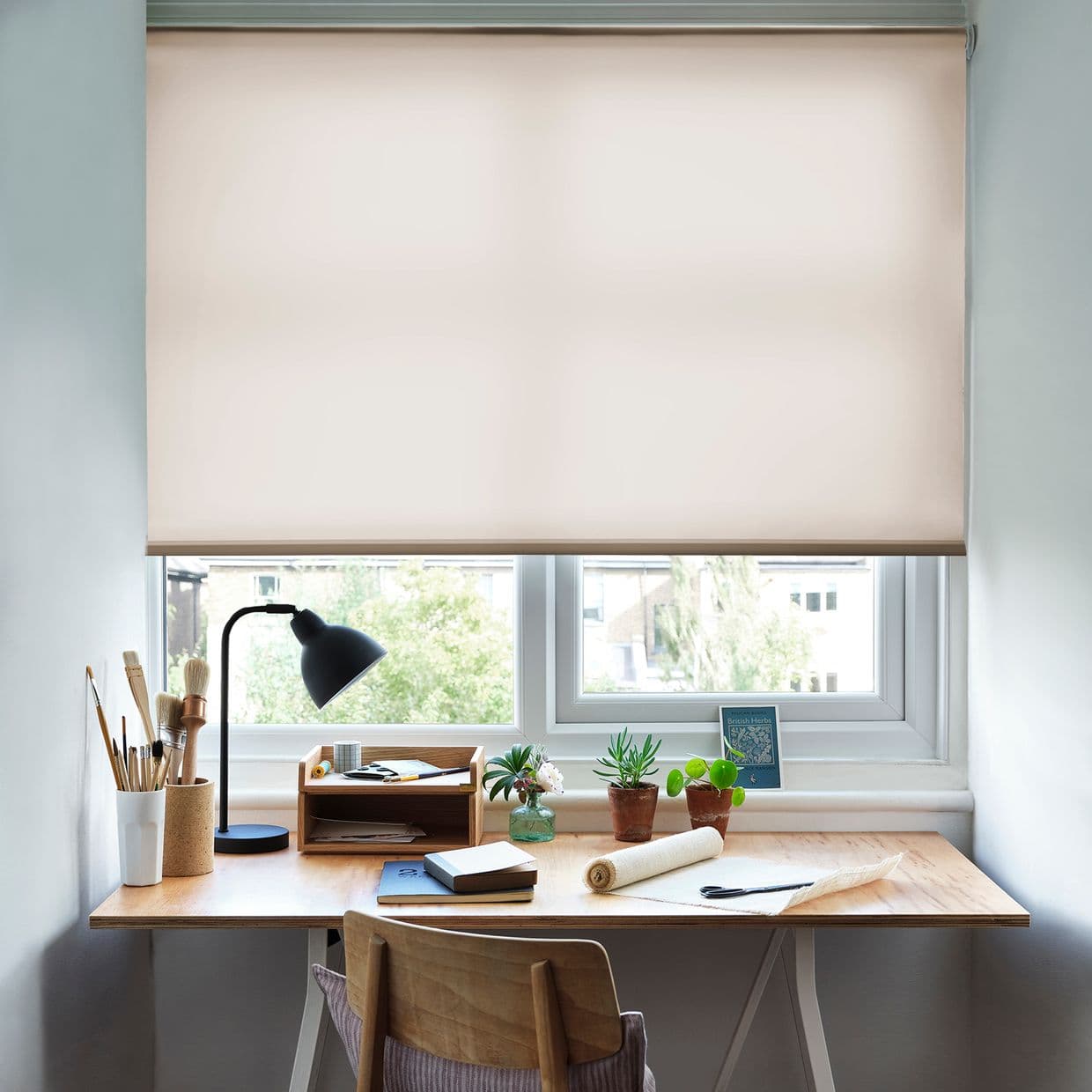 Object: Roller blind â€” beige, smooth fabric lowered over the window. Action: filtering soft daylight through a narrow gap at the sill. Context: tidy wooden desk workspace with lamp, brushes, potted plants, outside trees and buildings. British Herbs