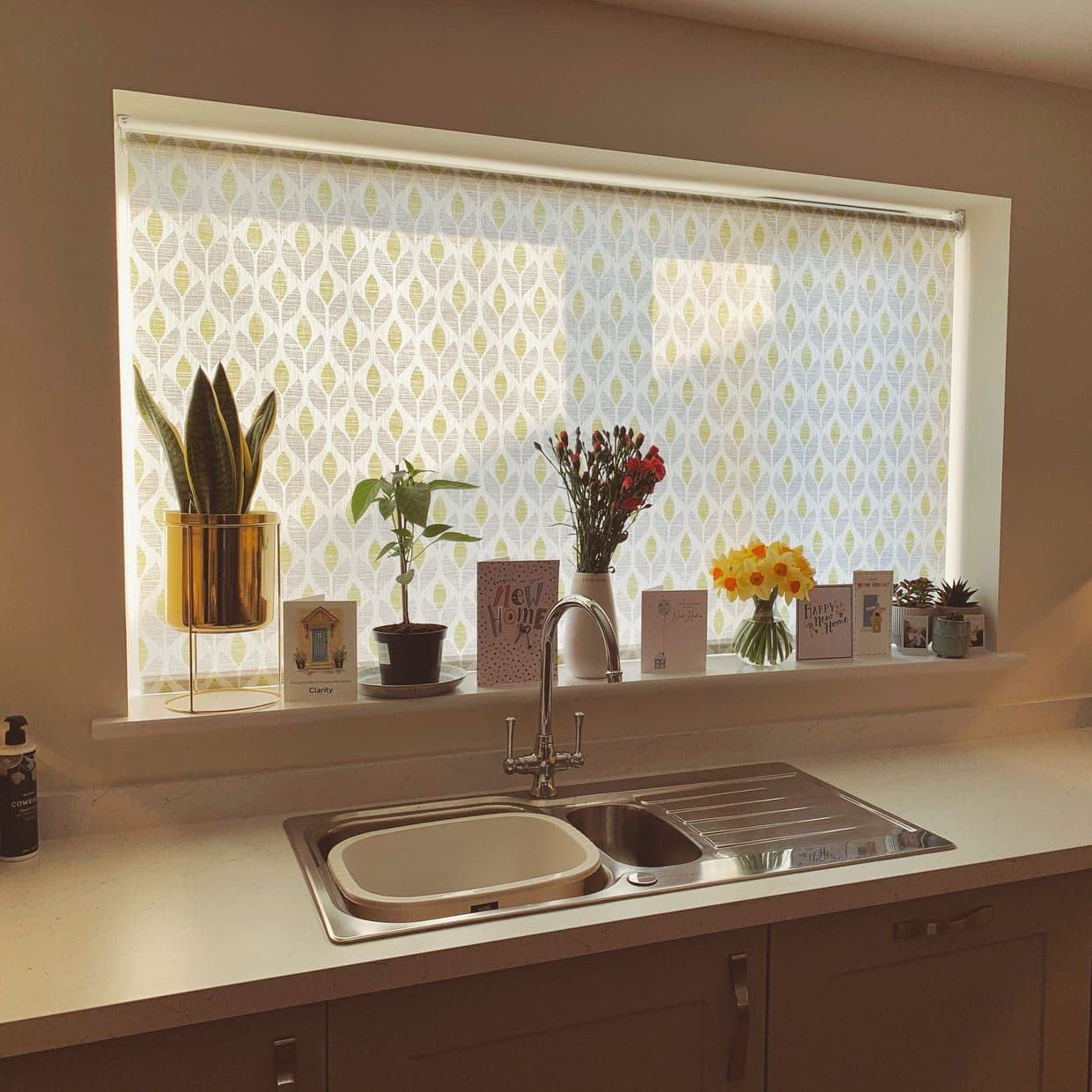 Patterned roller blind, lowered and semiâ€‘translucent, filtering daylight over a kitchen sink; windowsill holds potted plants, vases of flowers, and several greeting cards.

Clarity
New Home
Happy New Home
COWSHED