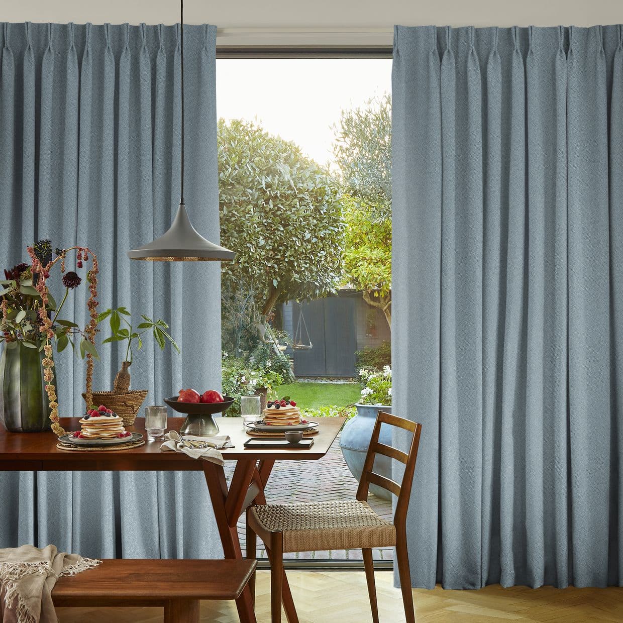 Full-length pleated pale blue curtains, hung on a rod and parted at center with soft folds; framing a sunlit garden; dining table, wooden chairs, pendant lamp and breakfast plates inside.