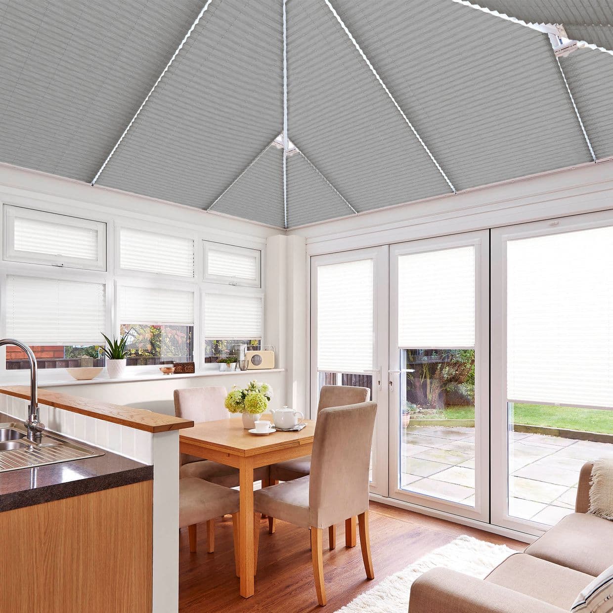 Pleated cellular blinds installed across the conservatory's pitched roof and covering windows and French doors, filtering bright daylight; interior shows wooden dining table with upholstered chairs, sink and garden view.