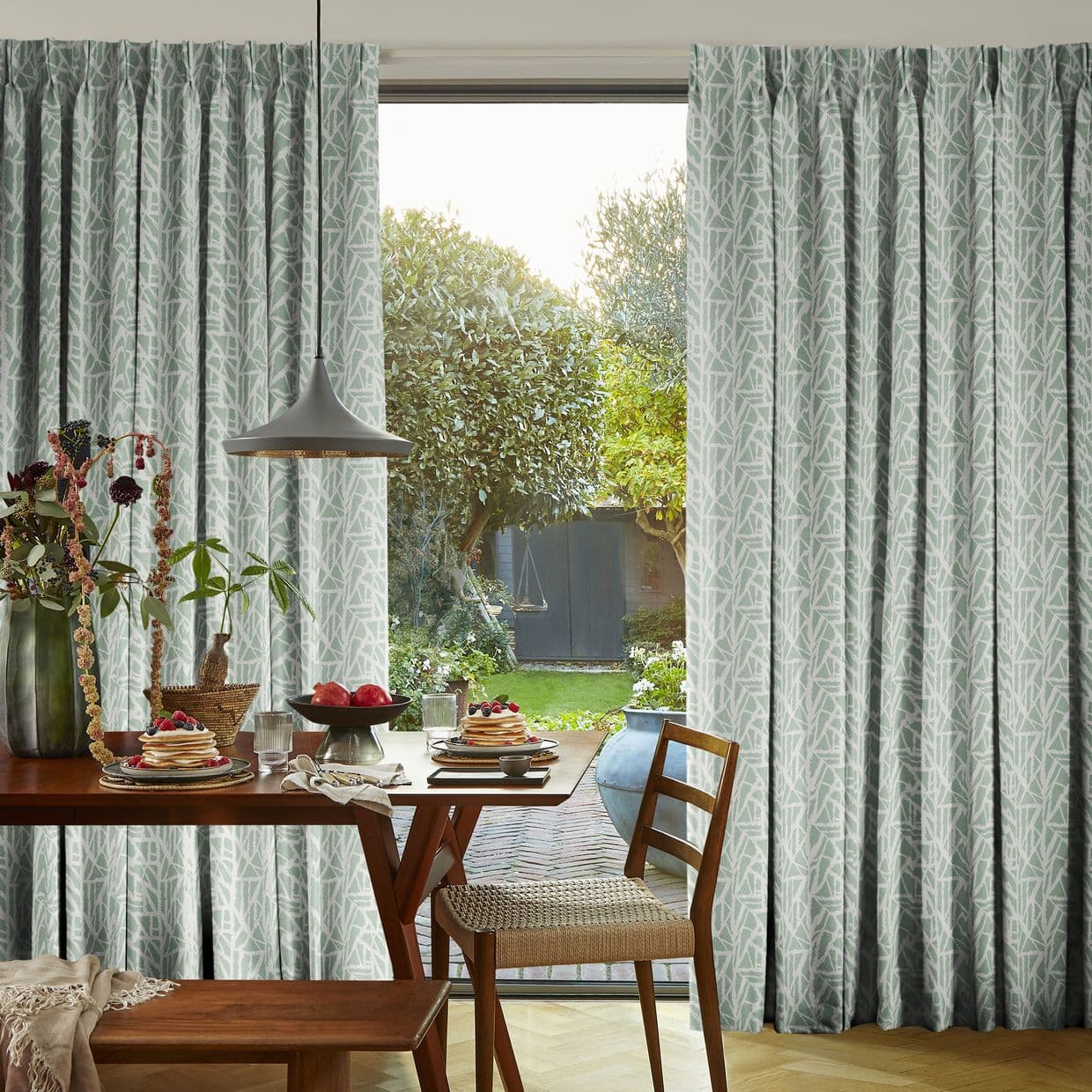 Floor-length patterned drapes (mint-green with white abstract twig motif) are mostly closed, parted centrally to frame the glass door. A breakfast table and pendant lamp sit inside; sunlit garden beyond.