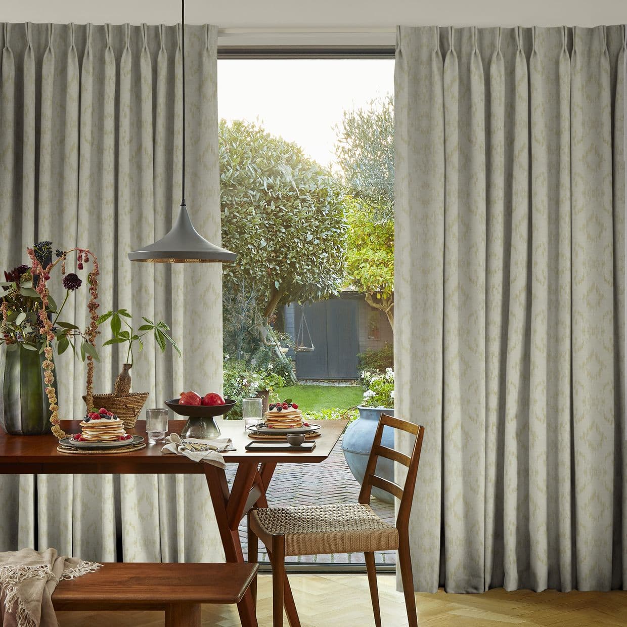 Pinch-pleat full-length curtains, pale patterned, hung floor-to-ceiling, parted centrally, gently filtering light; framing a dining table set for breakfast and a sunlit backyard with trees and a shed.