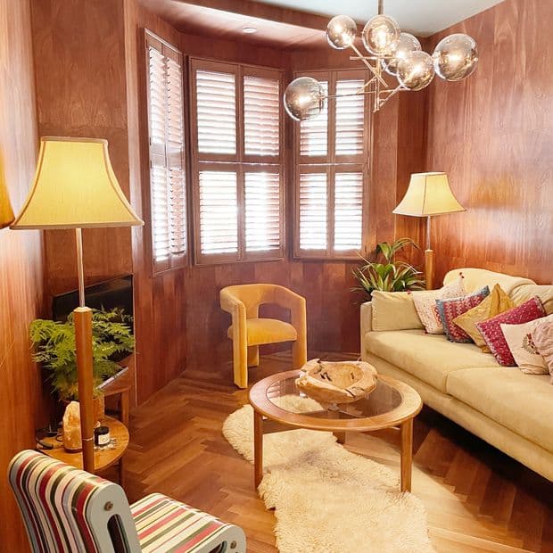 Wooden plantation shutters on a bay window, closed with angled louvers filtering soft daylight; set in a woodâ€‘paneled living room with warm lamps, beige sofa, round coffee table, and sheepskin rug.