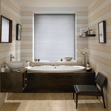 Horizontal venetian blinds lowered over the window, closed thin slats diffusing soft daylight; set above a dark-wood bathtub in a lit bathroom with striped walls, sink, shelf and chair.