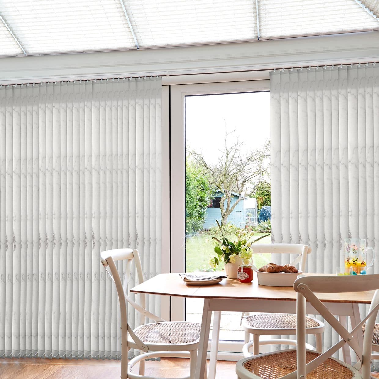 White vertical blinds with subtle marbled pattern across sliding patio doors, partially drawn and filtering diffuse daylight, dining nook with wooden table, chairs, vase and garden view.