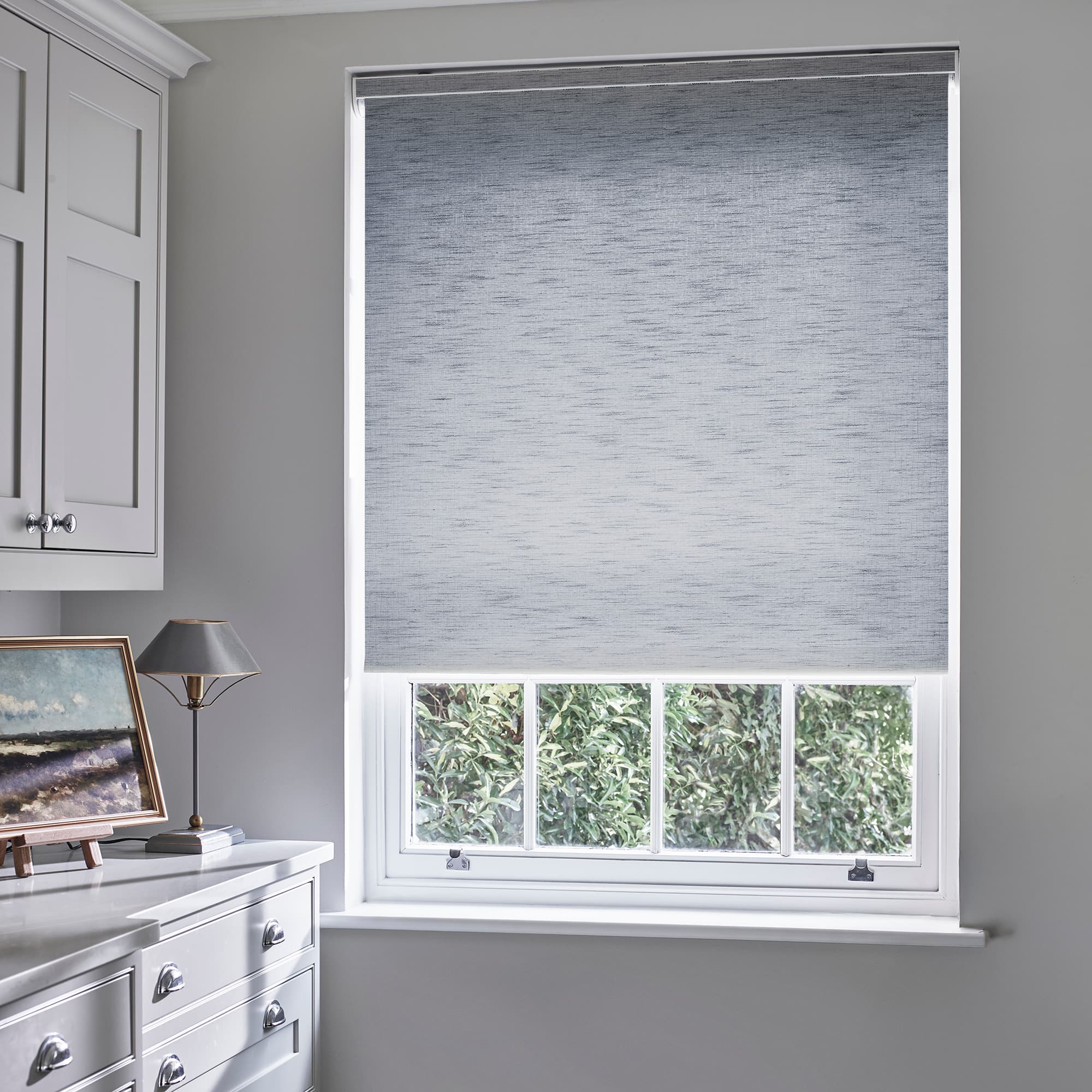 Grey textured roller blind lowered over upper two-thirds of a white sash window, partially closed and filtering daylight; pale-gray cabinetry, countertop with lamp and painting, hedge visible below.