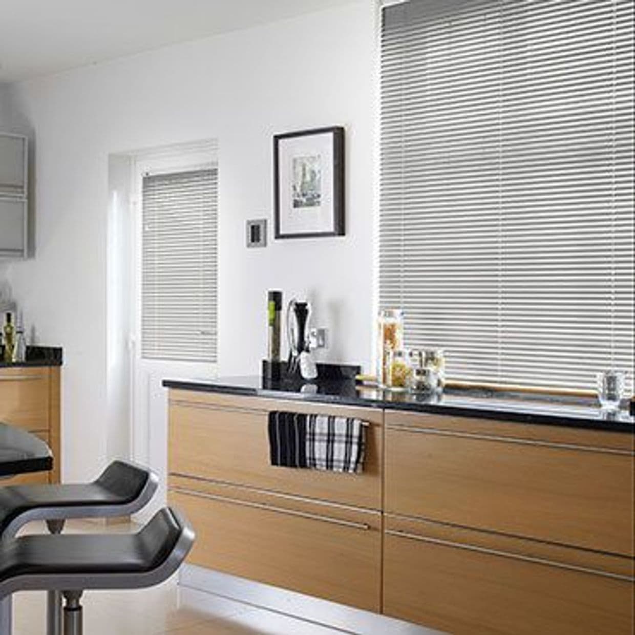 Horizontal metallic Venetian blinds covering a large window and door, fully closed and filtering soft daylight; context: modern kitchen with light-wood cabinets, black countertops, two bar stools, minimal decor.