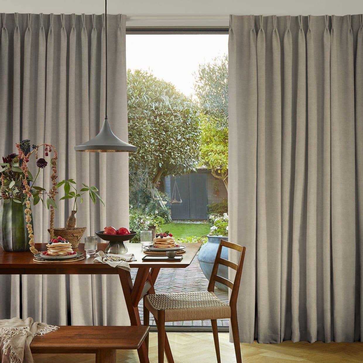 Floor-length, pinch-pleat grey curtains, drawn open at the center and hanging straight, framing a sunlit backyard; a wooden dining table set for breakfast with pancakes and a pendant light in foreground.