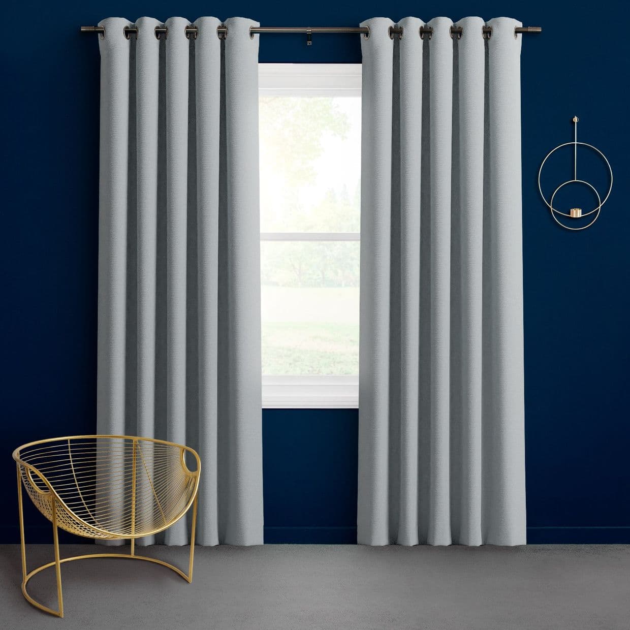 Light-gray grommet-top curtains, hanging in neat pleats on a metal rod, parted to reveal a sunlit window; navy-blue wall, minimalist gold wire chair and circular wall sconce.