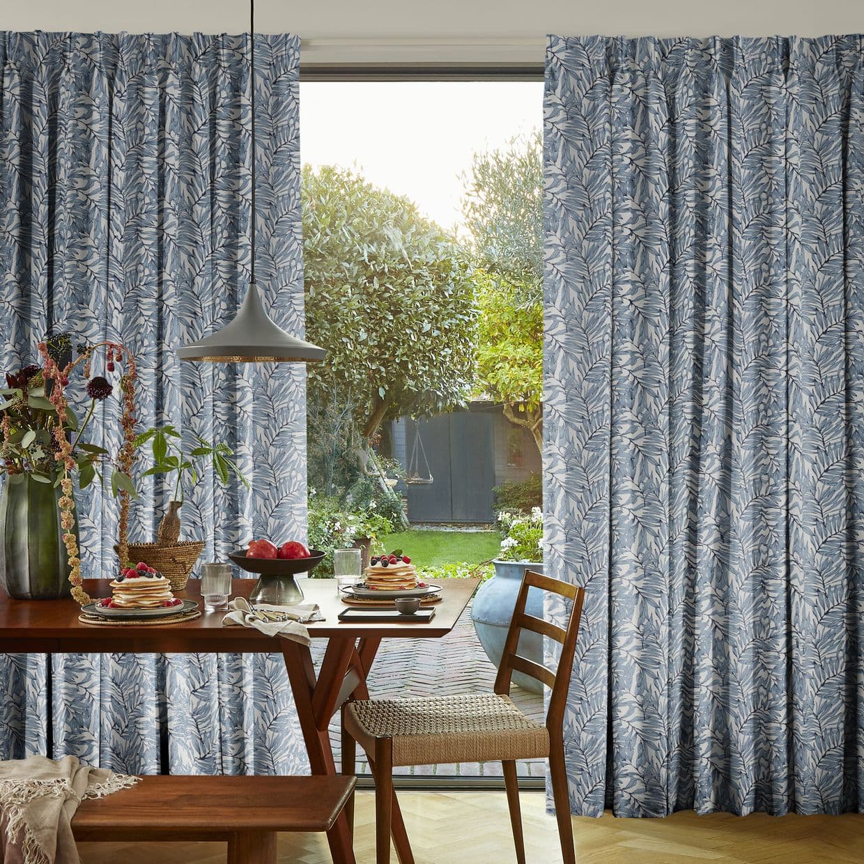Blue leaf-patterned pleated curtains, drawn aside to reveal a sliding glass door; filtering soft daylight over a wooden dining table set for breakfast, a pendant lamp, and a green garden view.