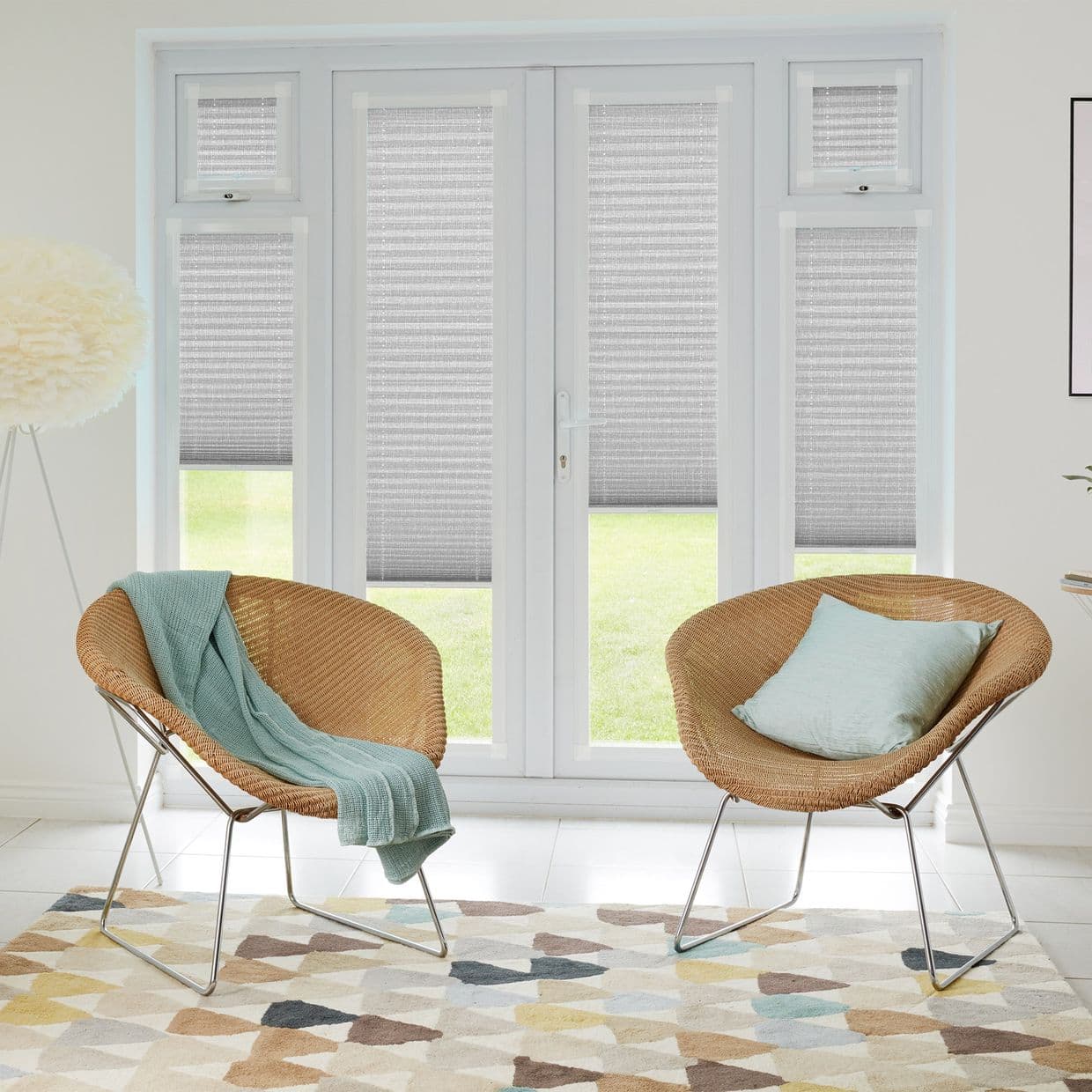 Pleated gray cellular blinds covering French doors and sidelights, partially lowered and filtering daylight into a bright sitting room with two wicker chairs, mint textiles, patterned rug.