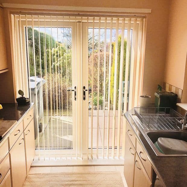 Cream vertical blinds cover glass patio doors, narrow slats hanging mostly closed and filtering sunlight; kitchen foreground with countertops, sink and dish rack, leading to sunlit garden outside.