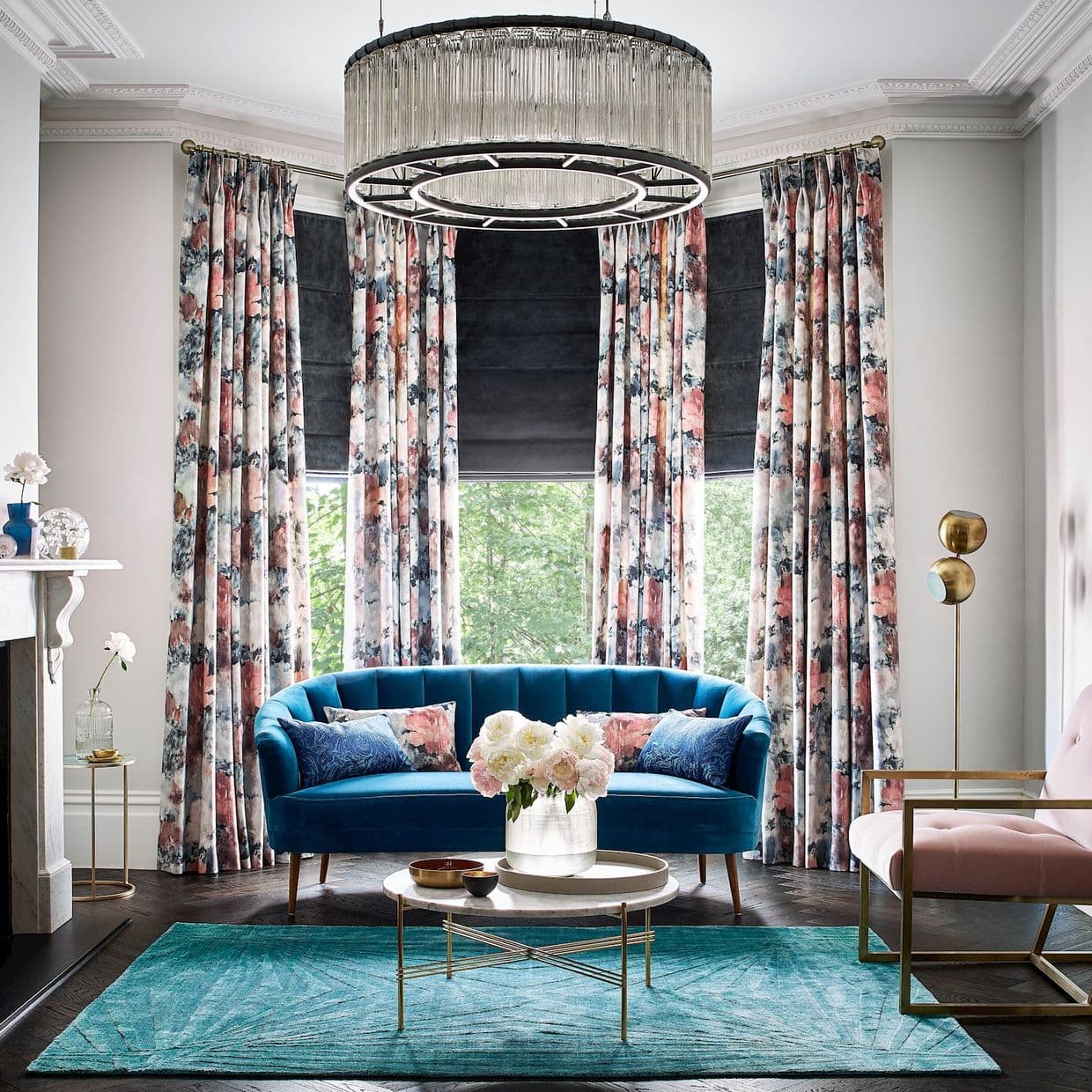 Floral patterned floor-length curtains on a brass rod, pleated and framing bay windows with dark Roman shades partially lowered, filtering daylight; elegant living room with teal sofa, chandelier, leafy view.