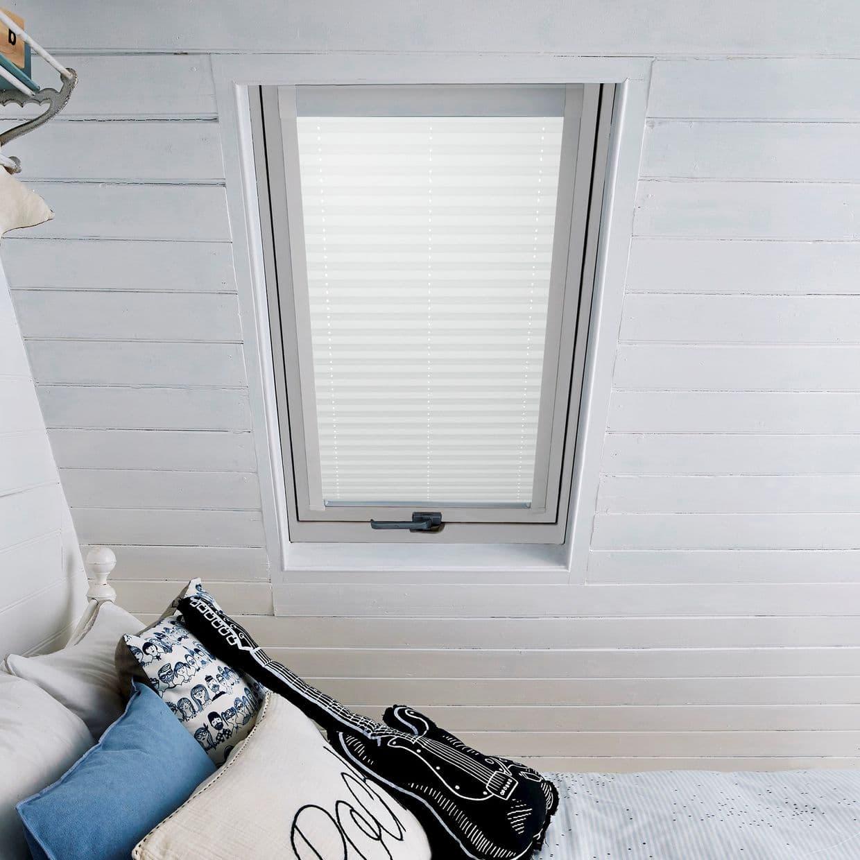 White pleated blind in skylight, fully closed with faint horizontal slats, filtering daylight into an attic bedroom with white paneled ceiling and a bed with blue, patterned and guitar-shaped cushions.