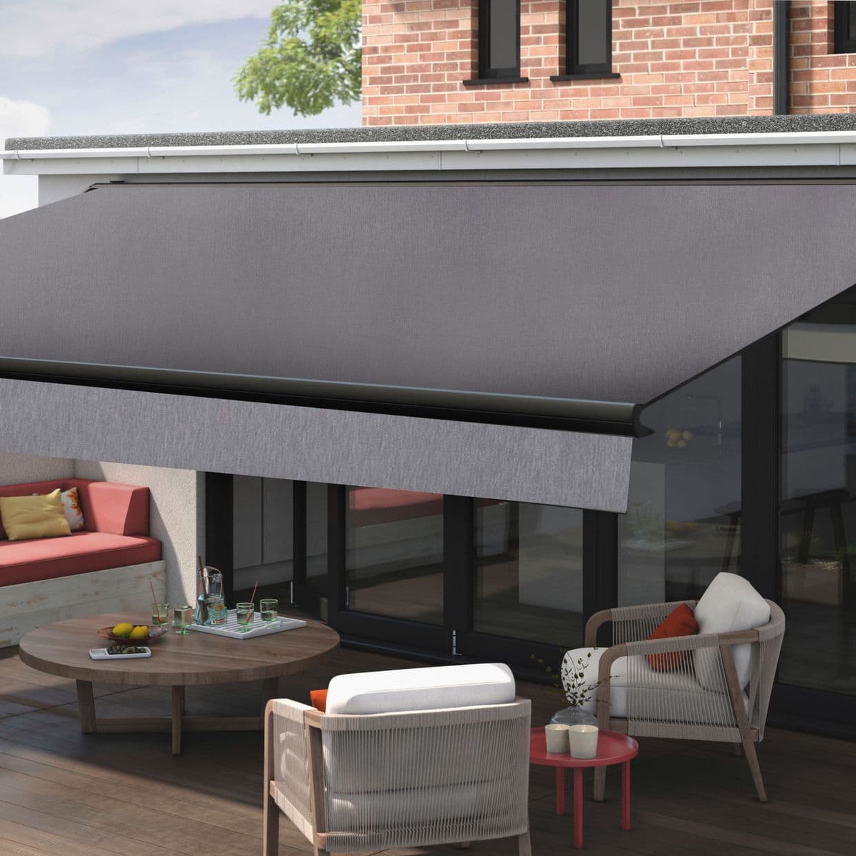 Large grey retractable awning with valance mounted at roofline; extended, shading and filtering light; over a patio with cushioned chairs, round wooden table, glass doors and brick upper wall.
