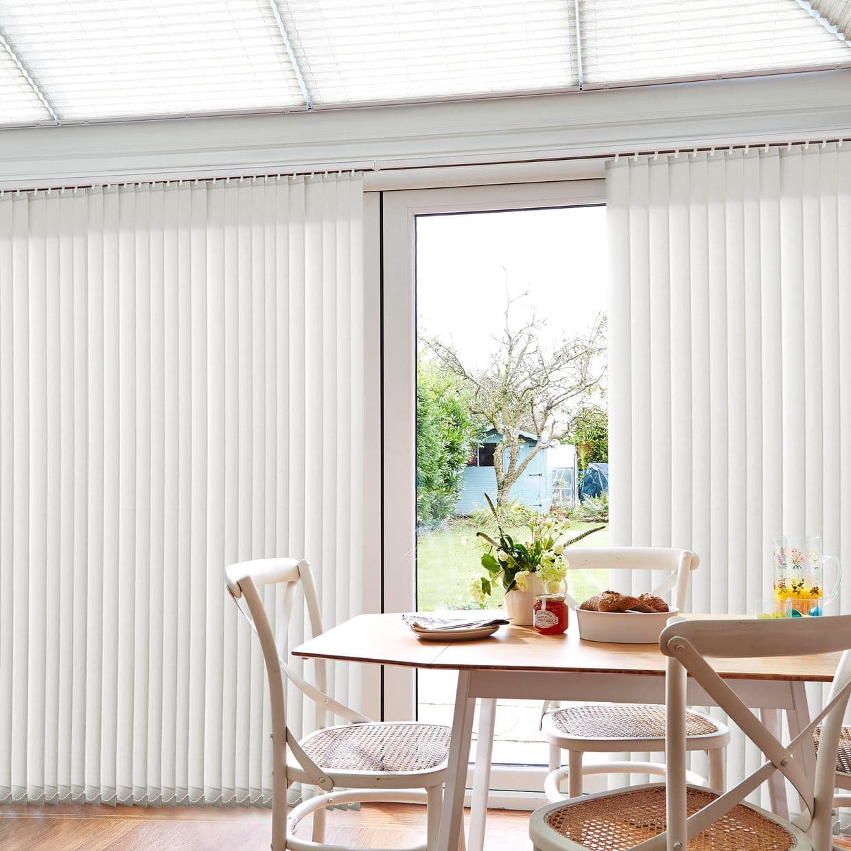 White vertical blinds, floor-to-ceiling, parted at center; filtering daylight and partially open to a glass sliding door; sunlit dining area with wooden table, chairs, and garden view.