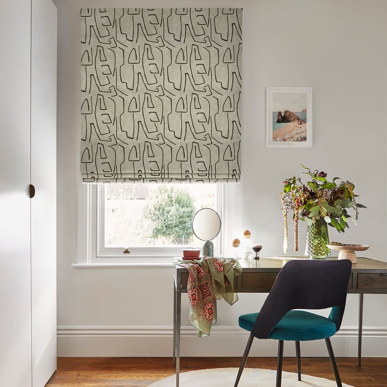 Roman blindâ€”beige fabric with bold black abstract-line patternâ€”lowered over a window, filtering daylight; small desk and teal chair beneath, vase of flowers, pale-walled home office.