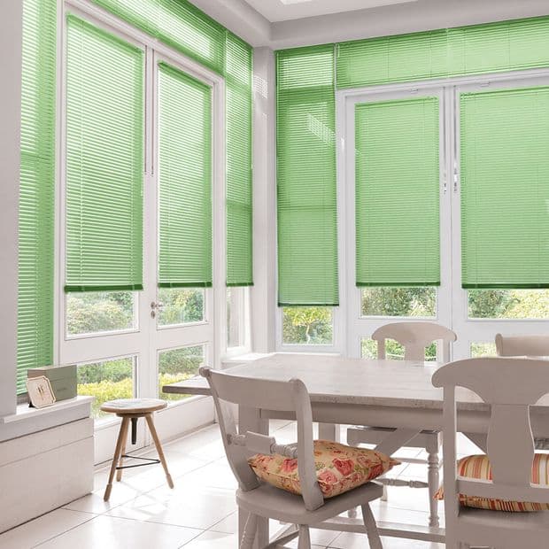 Green horizontal Venetian blinds covering tall corner windows and patio door, lowered with slats angled to filter light; bright dining sunroom with white table and chairs, cushions, and garden view.