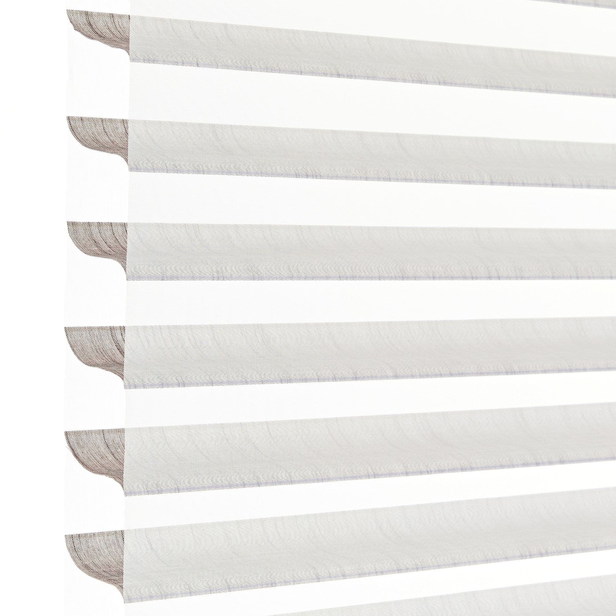 Sheer zebra roller blinds with alternating opaque and translucent horizontal bands, lowered to filter and diffuse bright daylight, shown close-up against a plain white background in soft light.