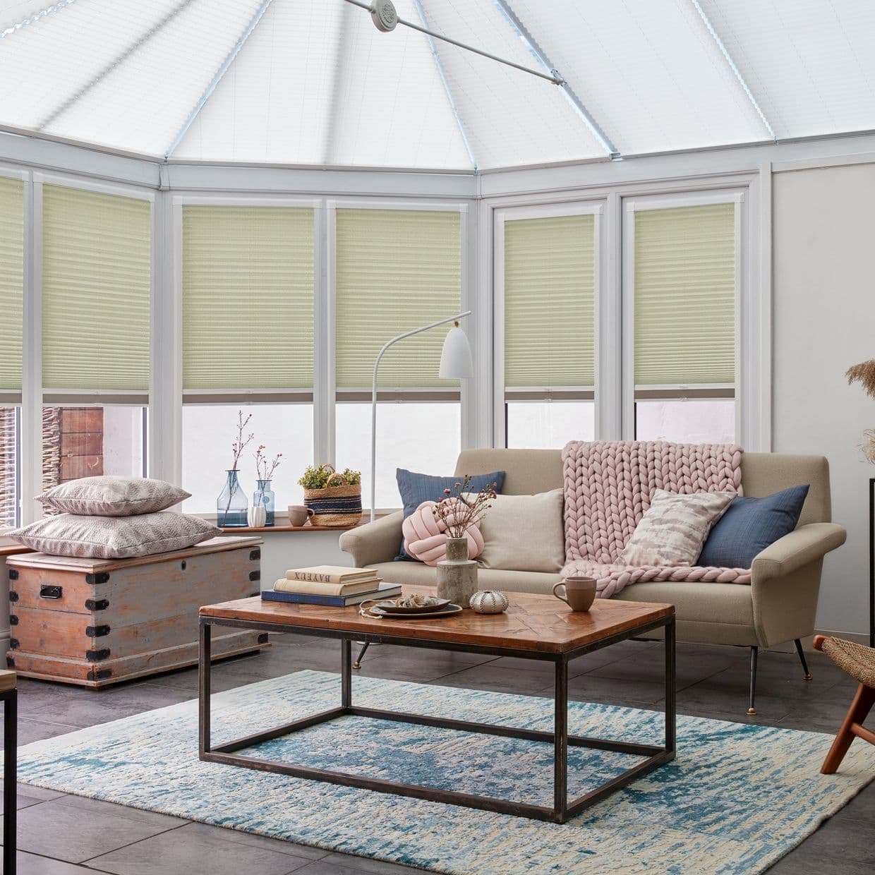 Pale sage pleated cellular blinds (covering tall conservatory windows, lowered with small lower gaps) â€” filtering soft daylight; a living room with beige sofa, wooden coffee table, and blue patterned rug.