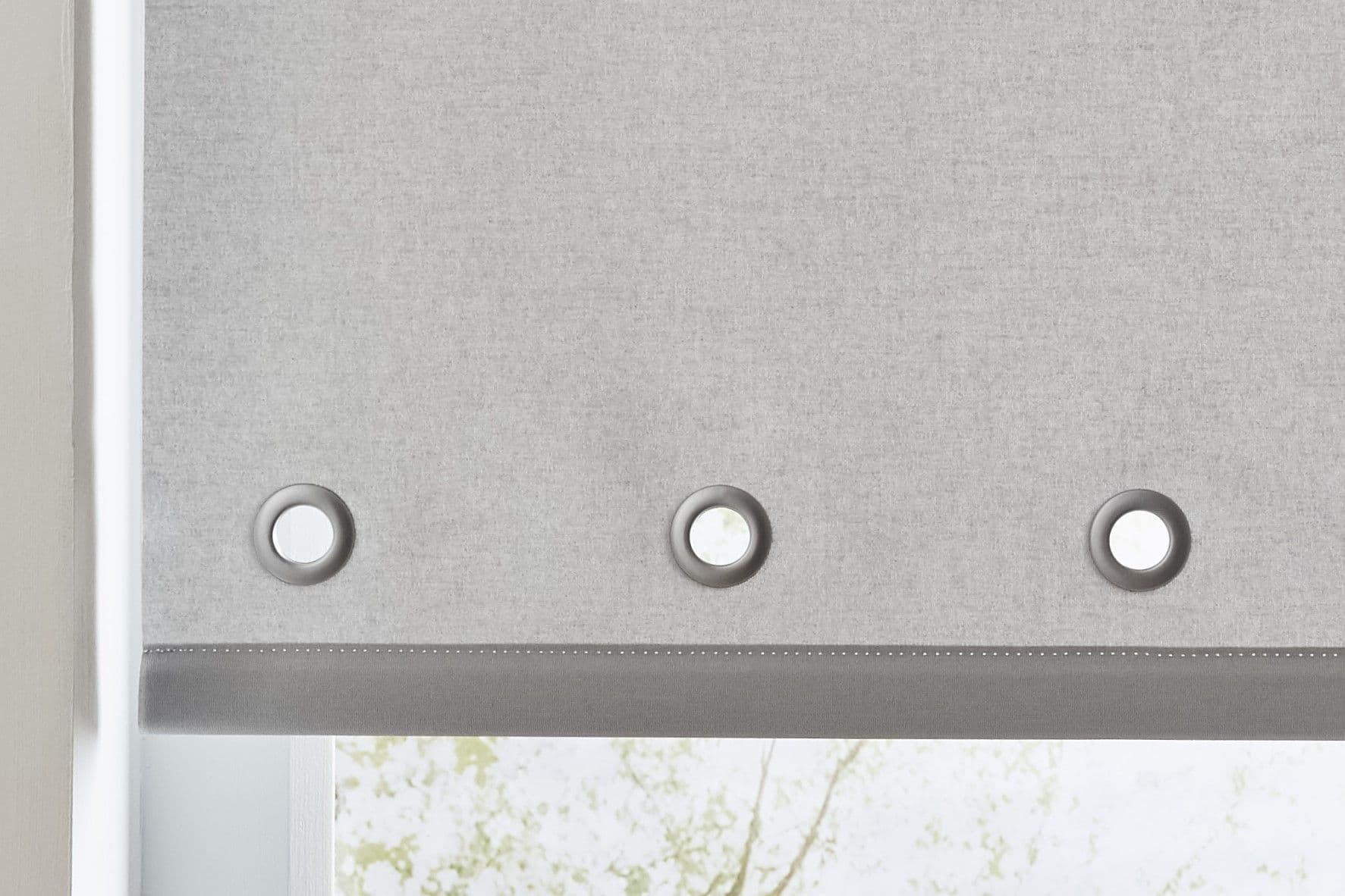 Grey fabric roller blind with three circular metal grommets, lowered and blocking most light while diffusing daylight from a window set in a white wall with a faint tree view.