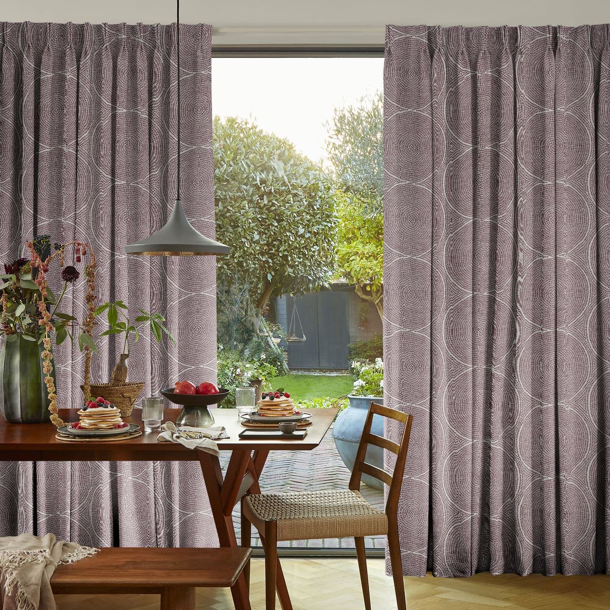 Floor-length, pleated mauve curtains with circular pattern, parted at center to reveal the glass door and frame sunlight; foreground dining table set for breakfast, pendant lamp, view of leafy garden.