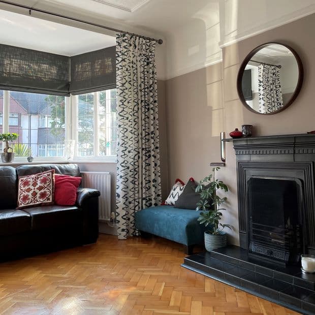 Patterned floor-length curtains (white with black squiggles) paired with lowered dark grey Roman blinds, filtering daylight at a bay window; cosy living room with leather sofa, fireplace, parquet floor.
