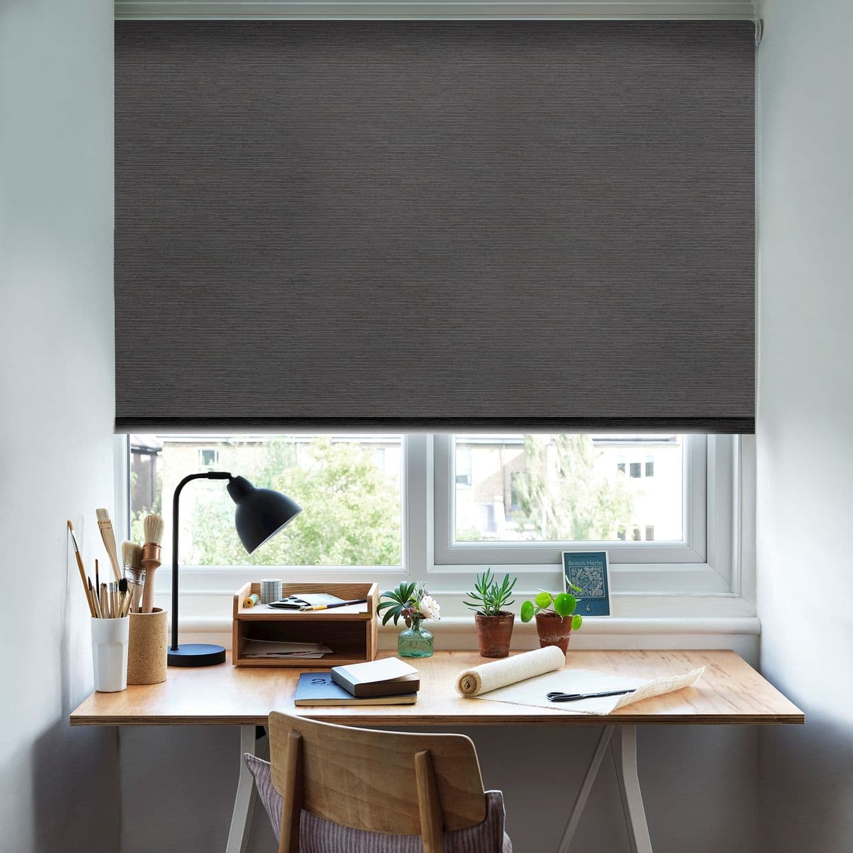 Dark grey fabric roller blind, lowered almost fully with a narrow bottom gap, blocking light; above a wooden desk holding a lamp, art supplies and potted plants in an office.