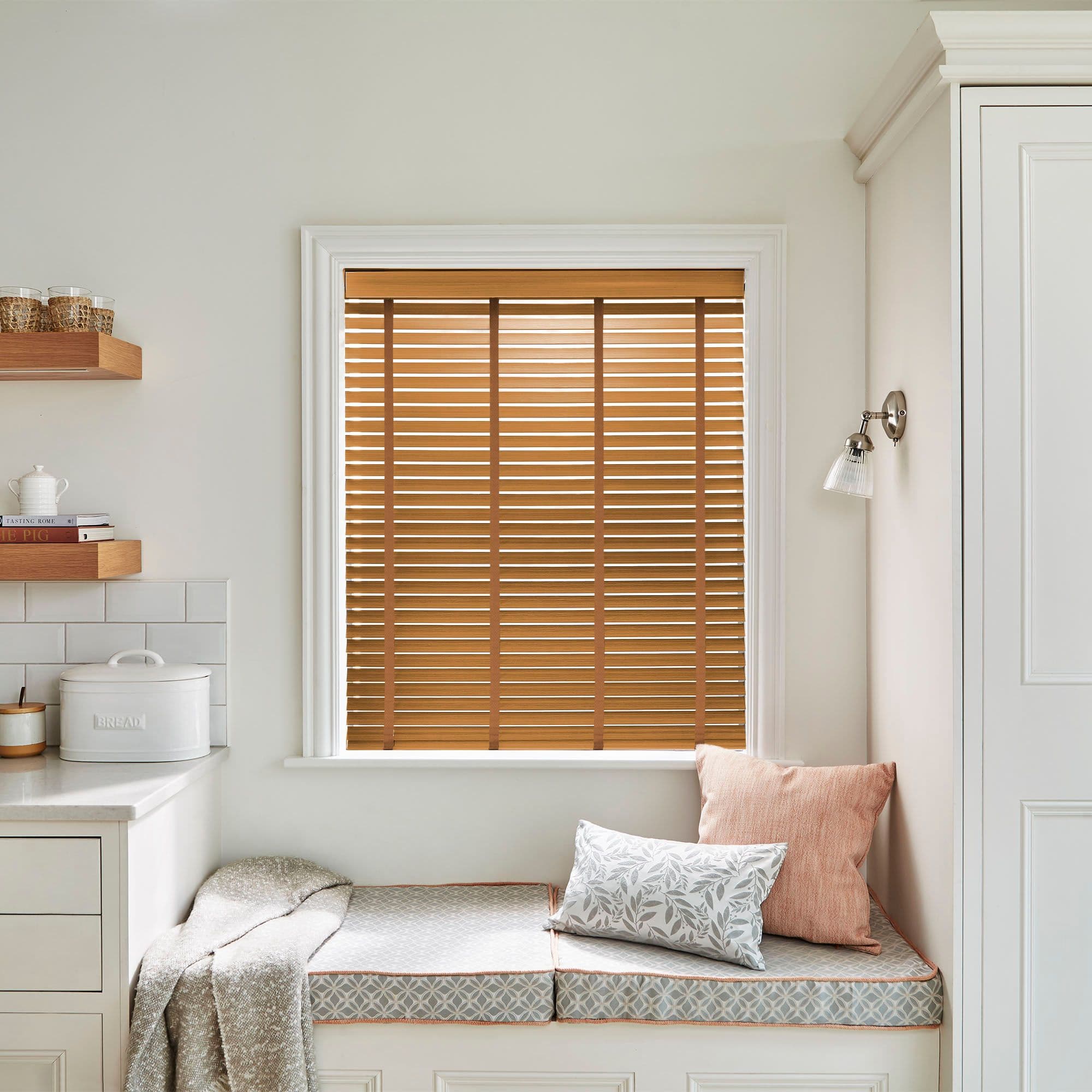 Wooden horizontal venetian blinds, centered and fully closed, filtering warm daylight through slats; kitchen window seat with pale cushions, woven throw, shelves and cabinetry in a bright, neutral nook. BREAD