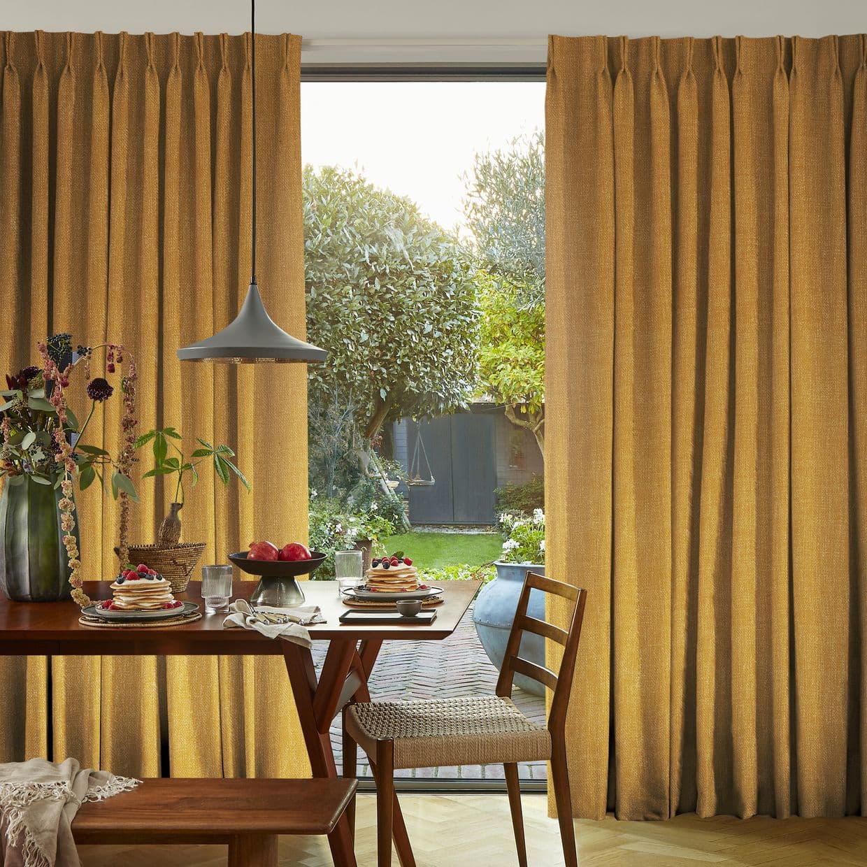Heavy textured mustard pinch-pleat curtains, parted centrally to frame a glass patio door and softly filter sunlight onto a wooden dining table set with pancakes, a vase, and a pendant lamp.