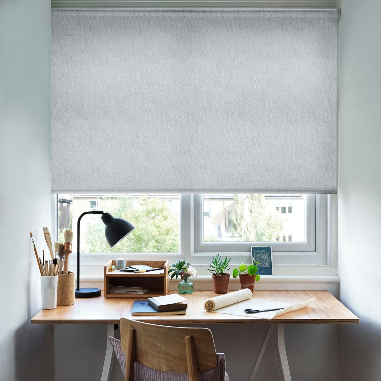 Light-gray textured roller blind fully lowered, gently filtering daylight; a wooden desk workspace beneath holds a lamp, paintbrushes, notebooks and small potted plants, with a leafy suburban view below.