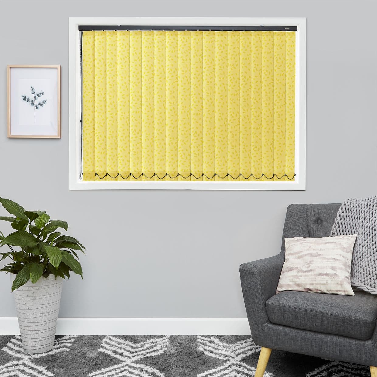 Yellow patterned vertical blinds covering the window â€” narrow slats with leaf print and scalloped beaded hem â€” fully closed, diffusing light in a gray living room with plant, armchair, framed print.