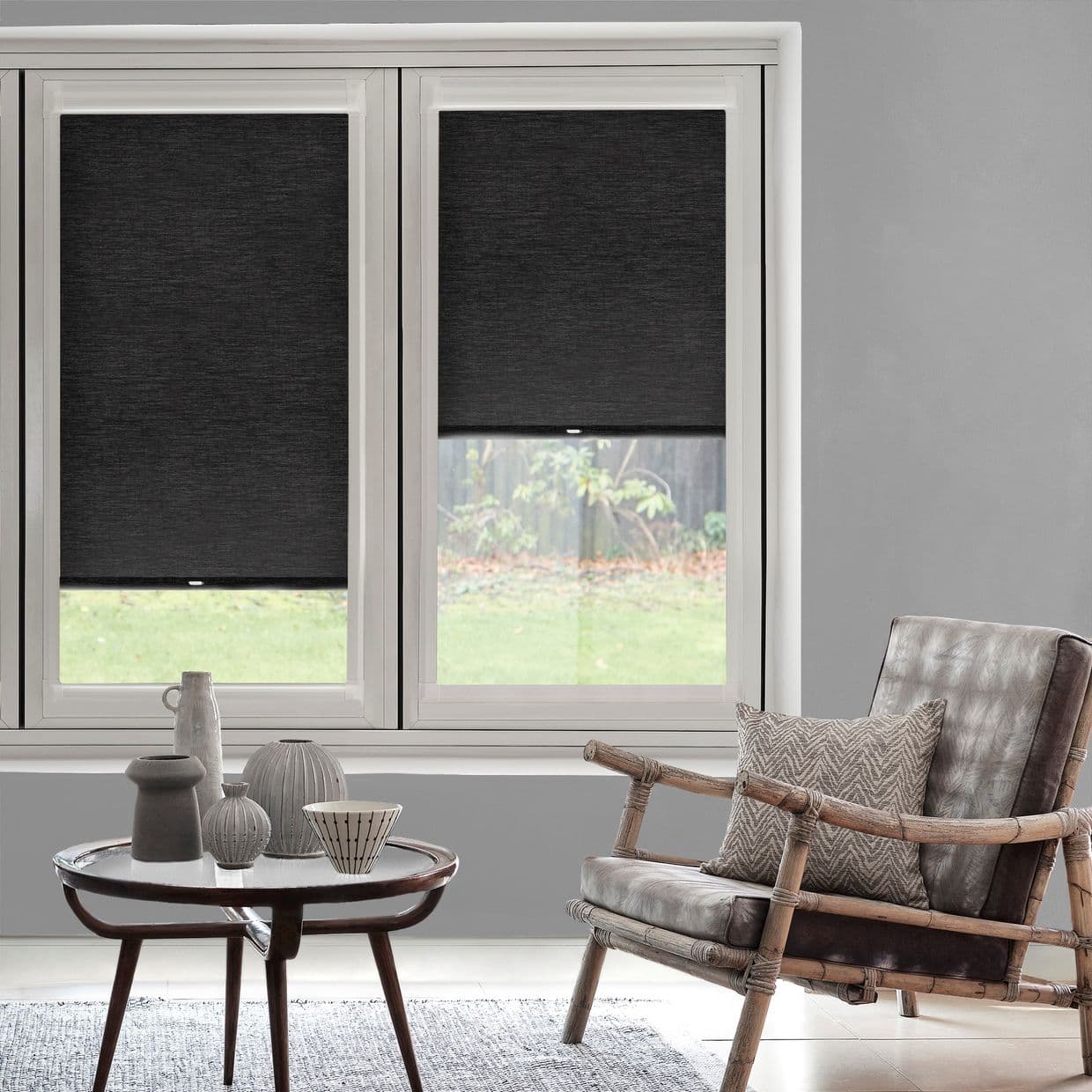 Charcoal roller blinds on two windows, mostly lowered with a narrow raised gap showing backyard; blocking light. Context: daylight, gray-walled living room with bamboo-framed leather chair and small coffee table.