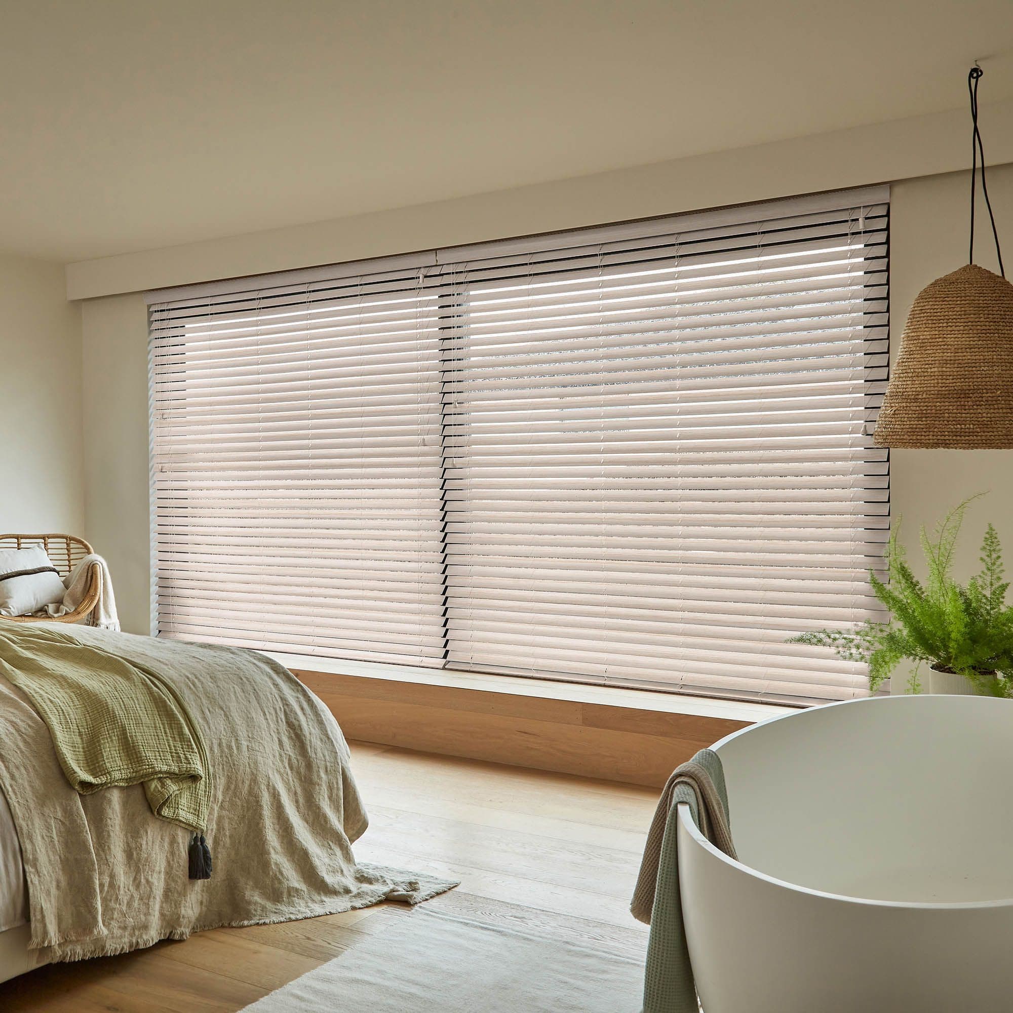 Horizontal Venetian blinds covering a wide bay window, slats closed and softly diffusing daylight; neutral, minimal bedroom with linen-covered bed, wooden floor, woven pendant light and a potted fern.