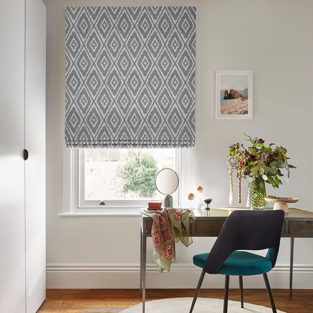 Grey-and-white geometric Roman blind lowered over a window, filtering soft daylight; small home office with wooden desk, teal chair, tabletop mirror and vase of flowers, pale walls and framed seascape.