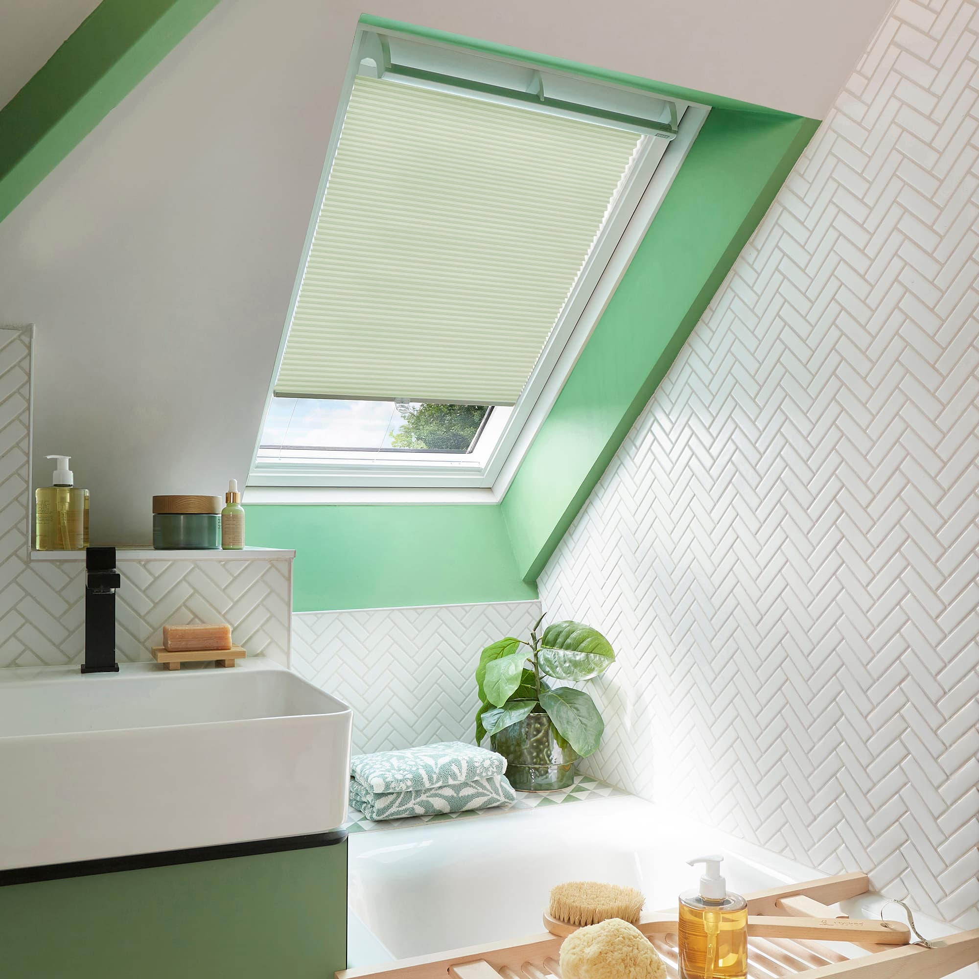 Light-cream pleated cellular blind fitted to an angled skylight, lowered and softly filtering daylight; set within a mint-green frame in an attic bathroom with white herringbone tiles, sink, tub, plant.