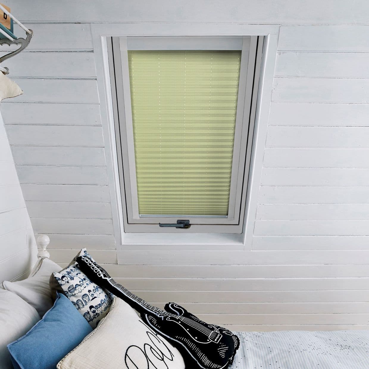 A pale green pleated blind, fully closed inside a white-framed skylight, filtering soft daylight; set in a sloped white shiplap attic ceiling above a bed with blue, patterned, guitar-shaped pillows.