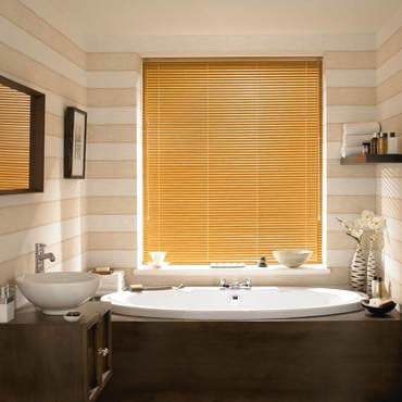 Golden wooden Venetian blinds covering a central window, fully closed and filtering warm daylight; set above a freestanding white bathtub in a contemporary bathroom with beige horizontal-striped walls and decor.
